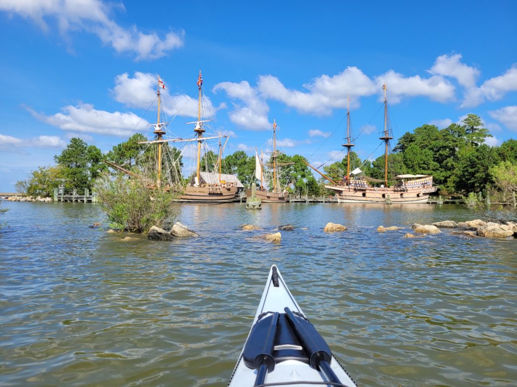 bow of a kayak in the foreground with large, 17th century replica ships in the background